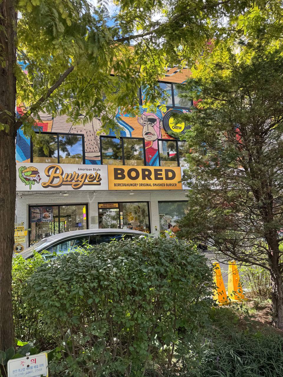 나무 사이로 보이는 Burger Bored 가게 외관, 노란 BORED 간판과 2층 벽면의 선글라스 낀 얼굴 벽화