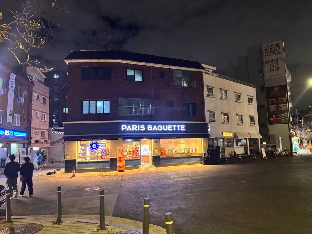불 켜진 빠리바게뜨 간판이 달린 붉은 벽돌 건물과 그 옆 흰 건물, 길가의 볼라드와 걸어가는 두 사람이 보이는 밤 풍경