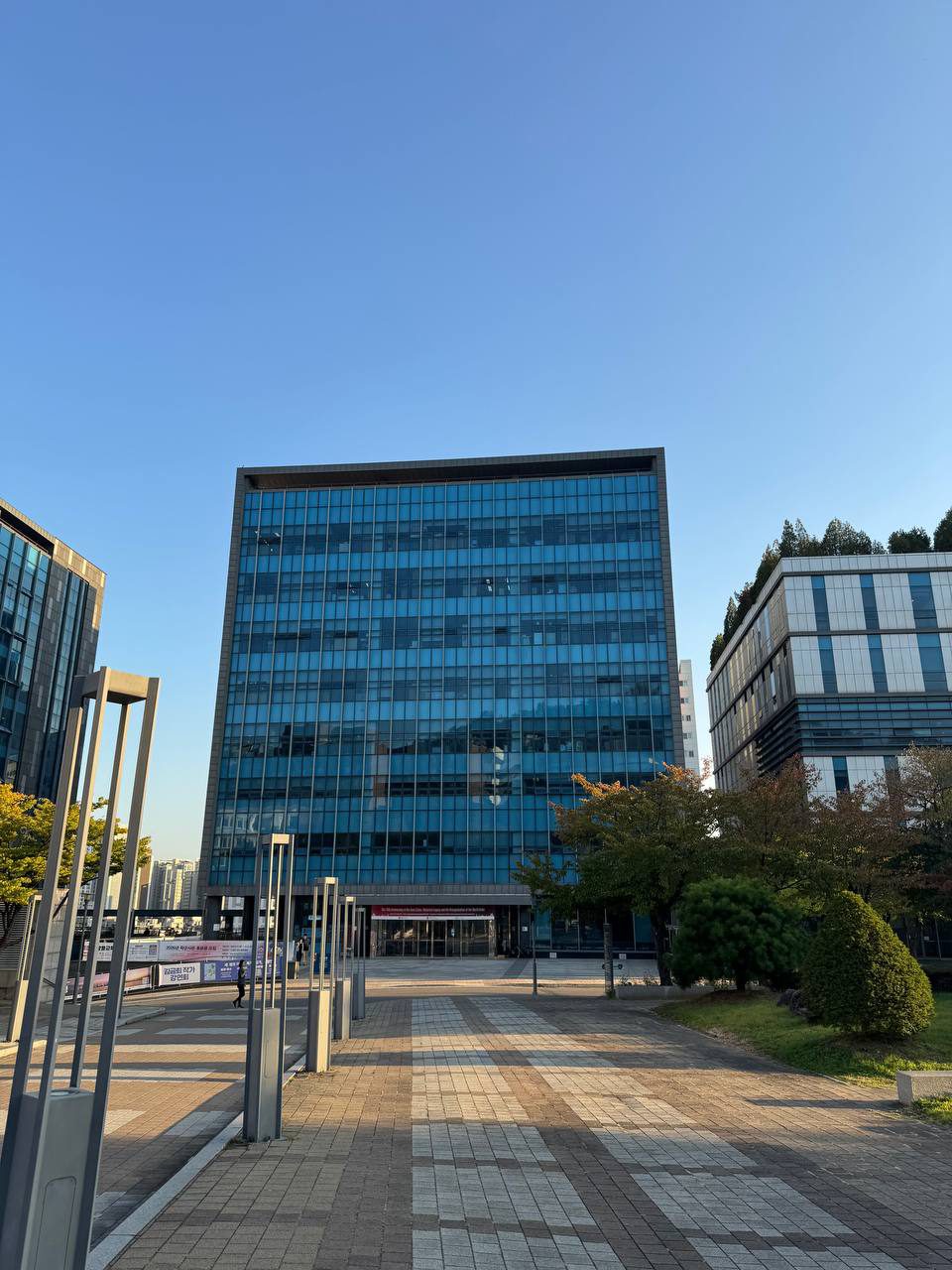 푸른 유리창이 격자 모양으로 이어진 건물 정면으로 보도블록 길이 나 있고, 왼쪽에는 회색 조명 기둥들이 줄지어 서 있다