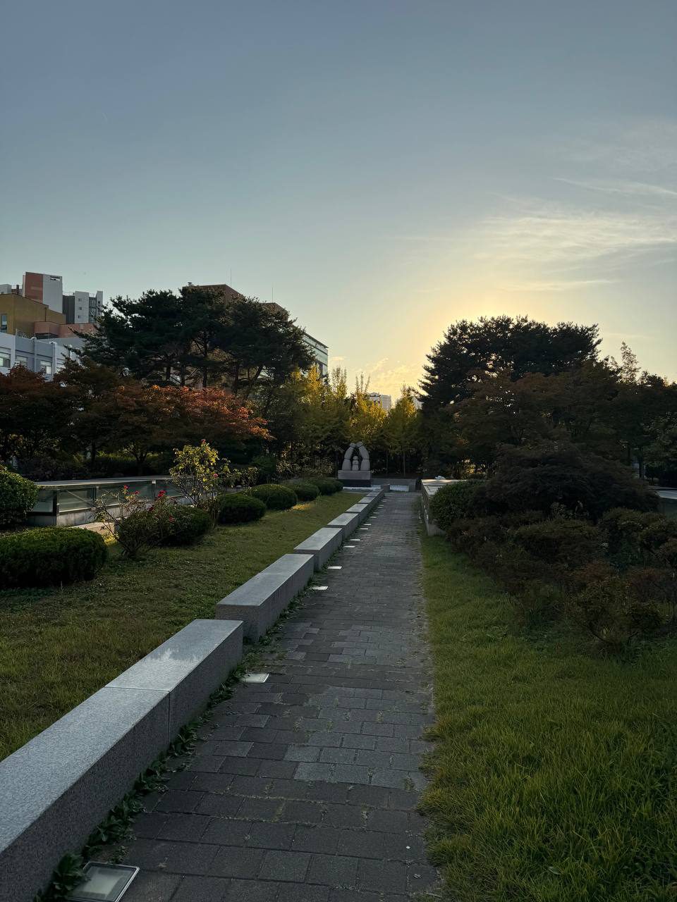 잔디와 수풀 사이로 좁은 벽돌길이 이어지고, 왼쪽에는 낮은 돌 벤치가, 길 끝에는 조형물과 소나무가 보인다