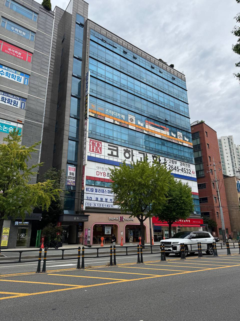 파란 수학학원, 붉은 국어학원 간판이 붙은 건물 옆으로 스터디카페와 과학전문 학원이 들어선 유리 외벽 고층 건물, 1층에 배스킨라빈스가 있는 거리