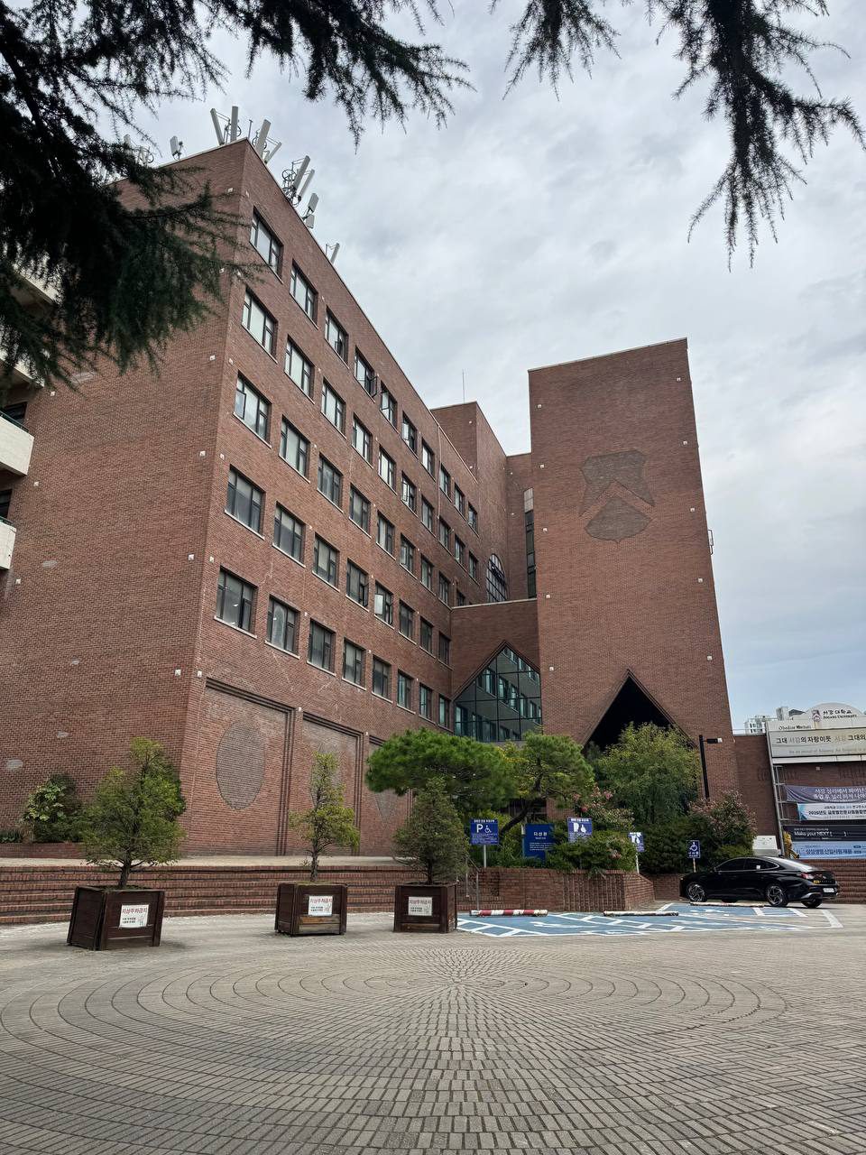 붉은 벽돌로 지어진 다산관의 전경, 삼각형 입구와 유리창 앞에 나무가 있고 광장 가장자리에는 주차 구역이 보인다