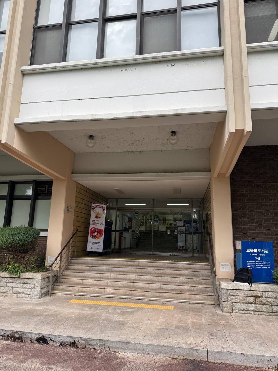 계단과 노란 점자블록이 놓인 로욜라도서관 1관 출입구, 오른쪽에는 파란 안내판이 있다
