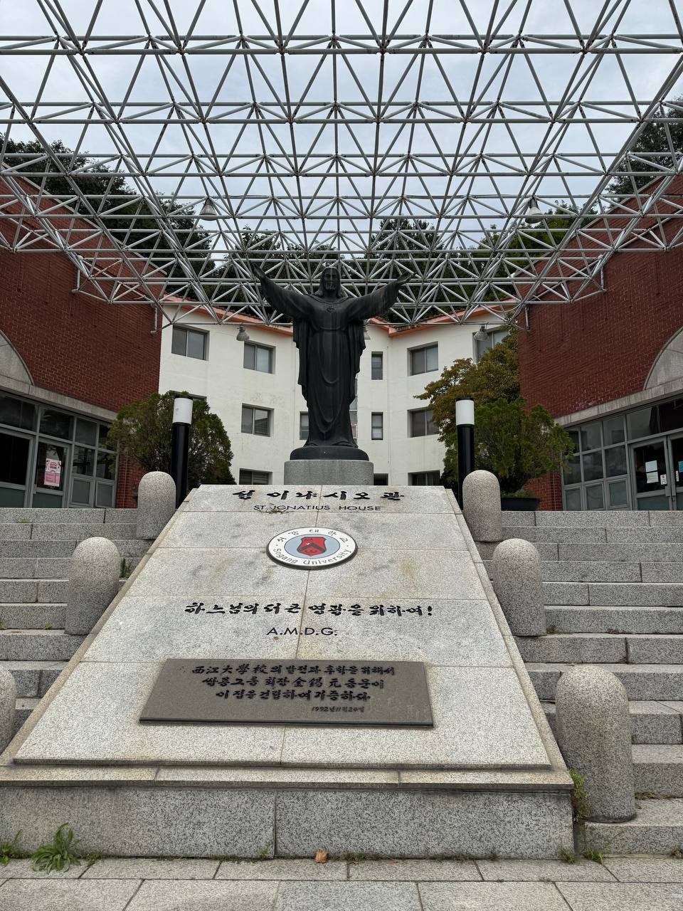 두 팔을 벌린 예수상 앞에 성 이냐시오관과 하느님의 더 큰 영광을 위하여라는 문구가 새겨진 돌이 놓여 있다