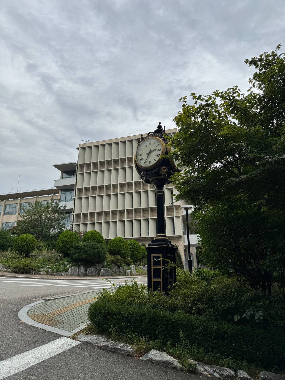 정면에서 올려다본 시계탑, 흰 바탕에 로마 숫자가 적힌 문자판과 위쪽 둥근 판의 글귀, 검은 기둥과 금색 선이 들어간 사각 받침대가 보인다