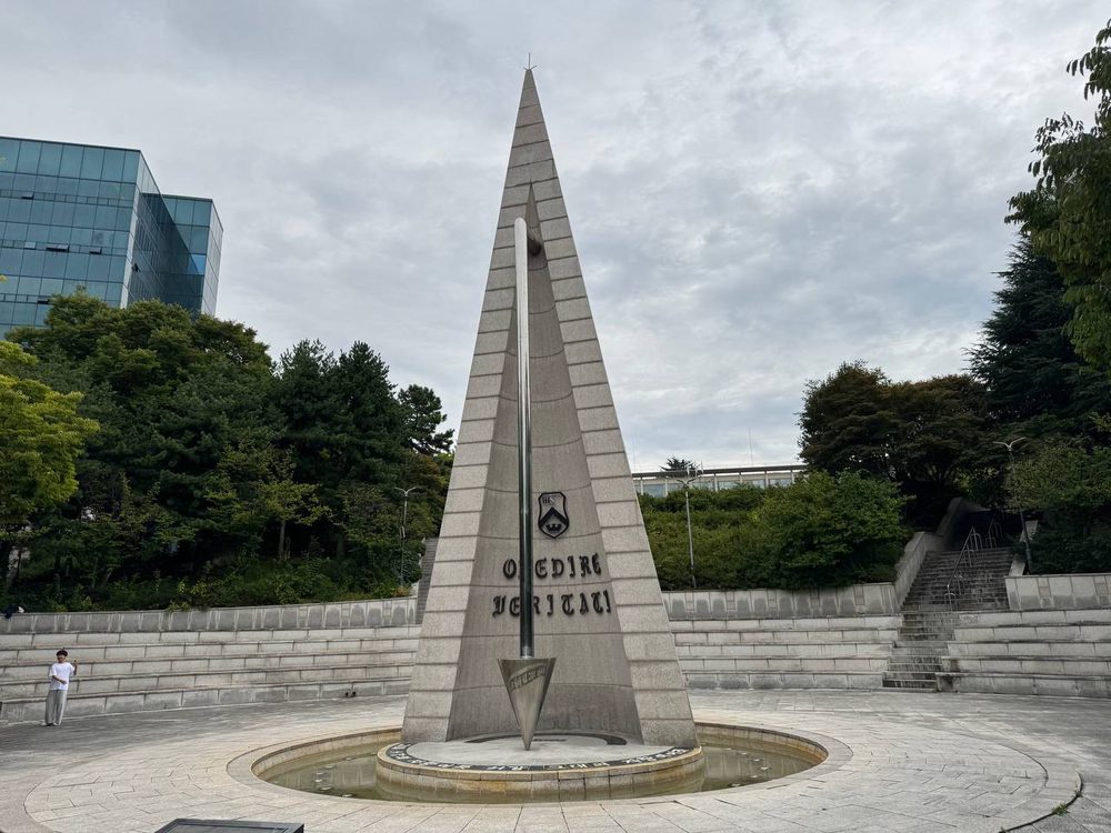넓게 잡은 알바트로스 탑과 광장, 왼쪽 석단 앞에서 한 사람이 사진을 찍고 있고 뒤로 유리 건물과 나무들이 보인다