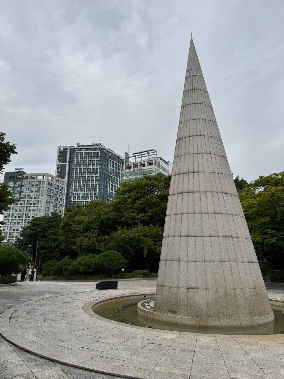 정문 쪽에서 본 알바트로스 탑의 뒷면, 가로줄 무늬가 들어간 화강암 원뿔 뒤로 고층 건물 세 동이 서 있다