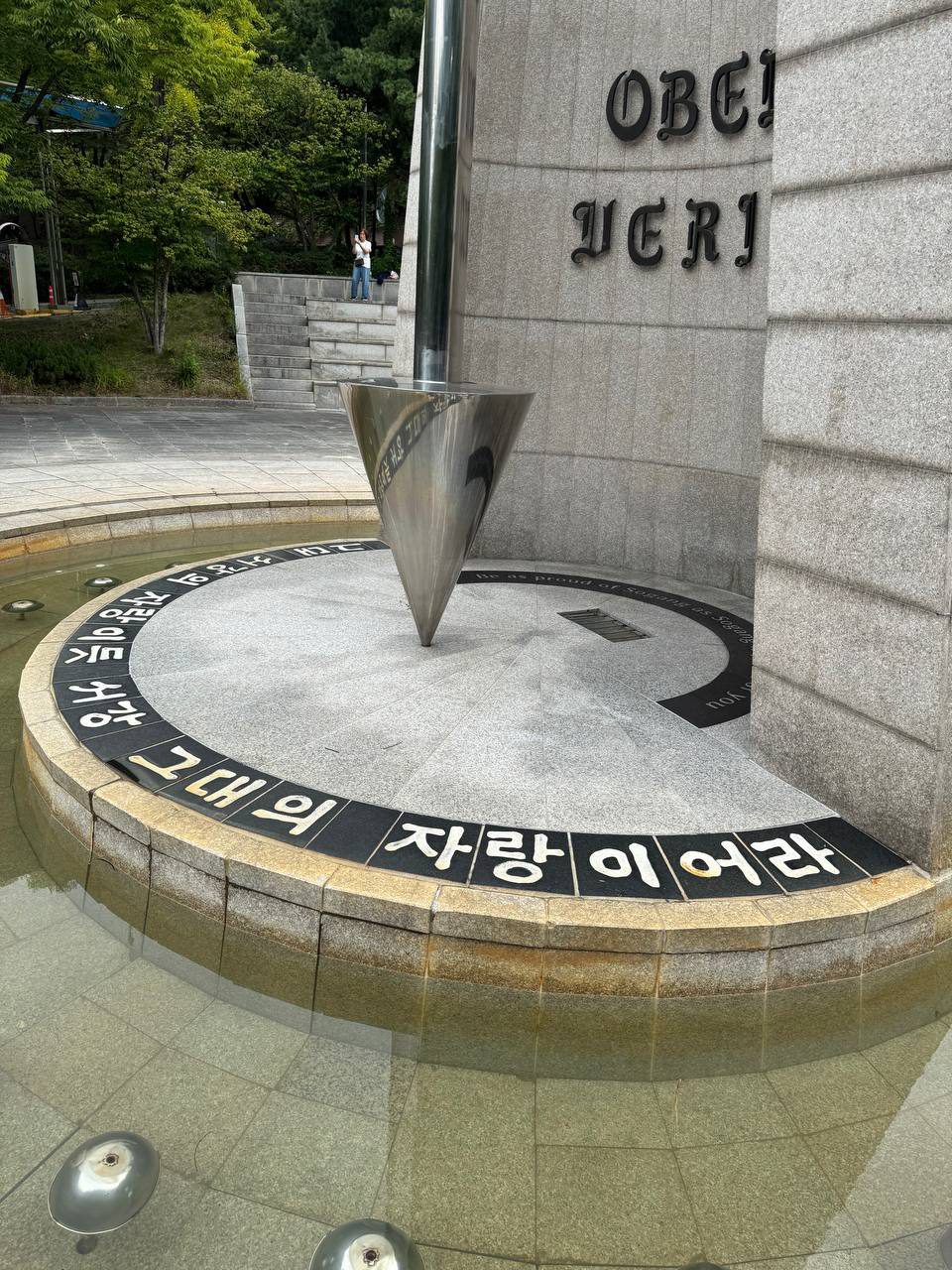 물이 찬 수반 너머로 가까이 찍은 바닥 글귀 ‘자랑이어라’와 영어 문구, 멀리 계단 위에서 한 사람이 사진을 찍고 있다