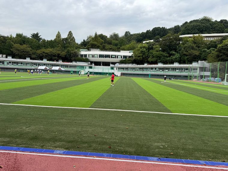 초록색 인조잔디가 줄무늬로 펼쳐진 서강대 대운동장에서 사람들이 축구를 하고, 건너편으로 엠마오관과 나무들이 보인다