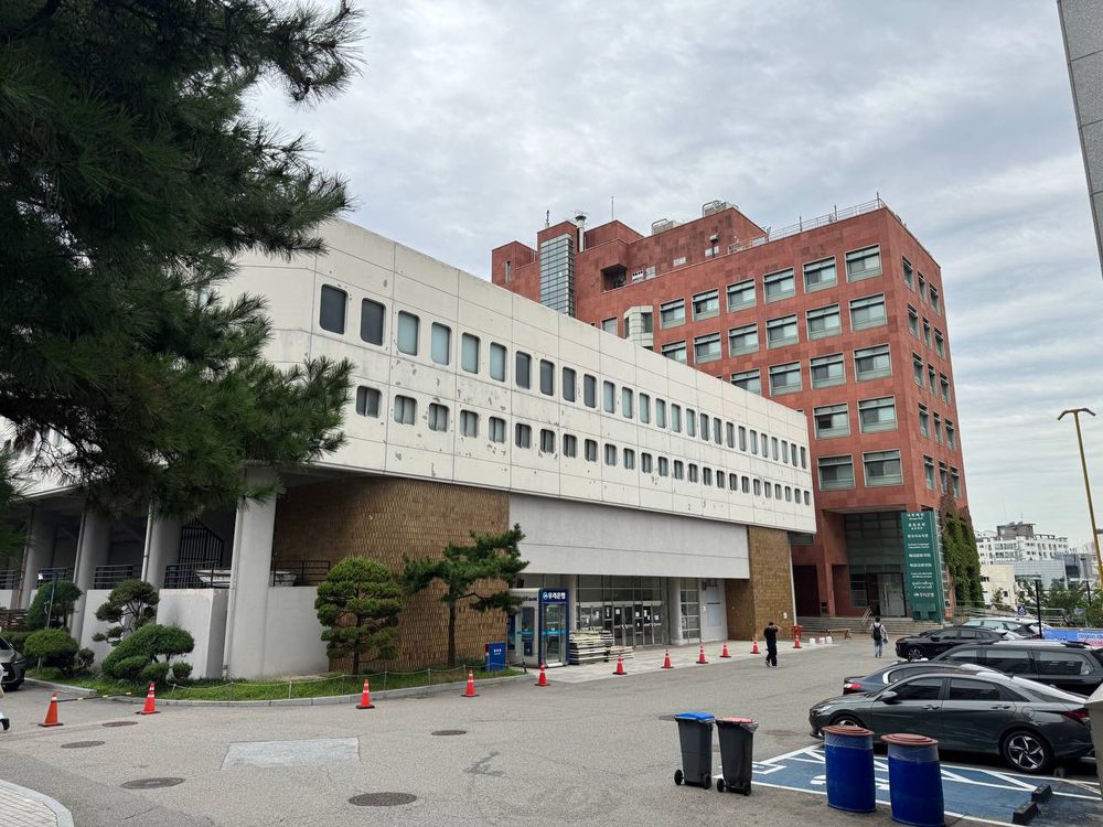 동그란 창이 줄지어 난 서강대 체육관의 흰 콘크리트 외벽과 그 뒤로 솟은 붉은 벽돌 건물, 앞쪽에 주차된 차들과 라바콘이 보인다