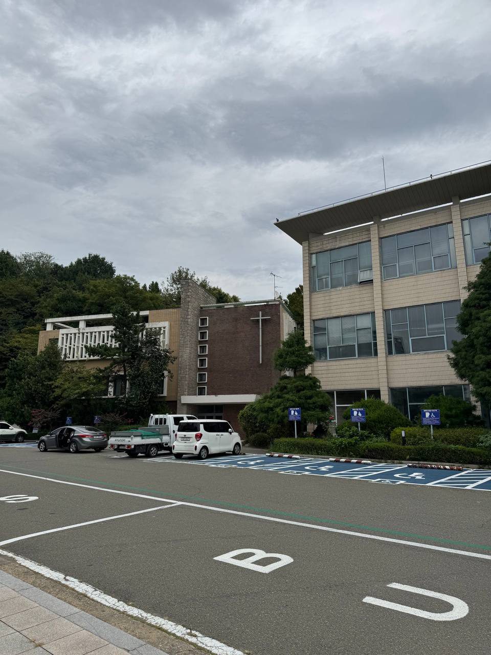 주차장 너머 나무 사이에 자리한 벽돌 사제관과 오른쪽의 큰 건물, 위로 흐린 하늘이 펼쳐져 있다