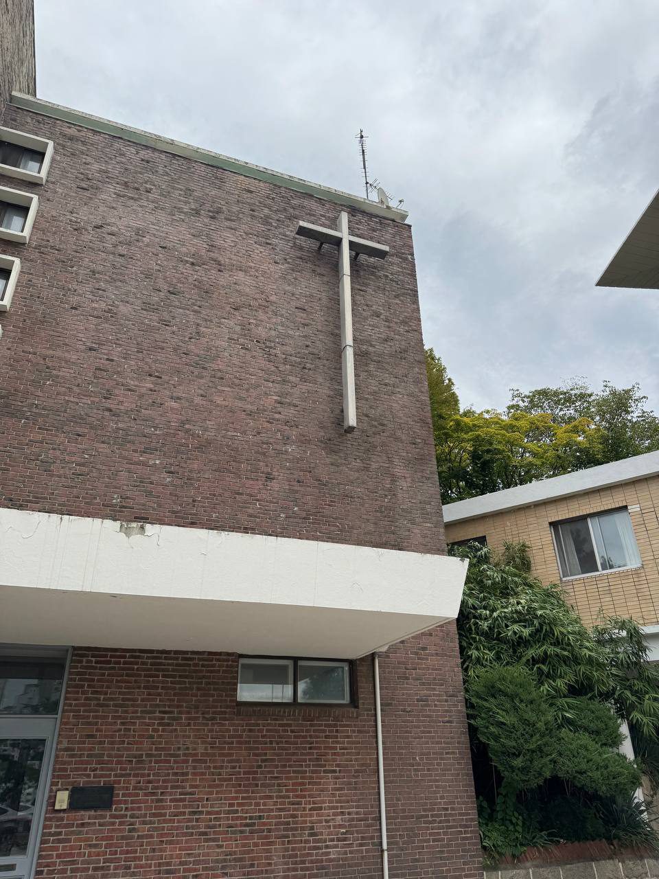 가까이서 올려다본 사제관의 벽돌 외벽과 긴 십자가, 아래쪽 흰 차양과 오른쪽의 무성한 나무