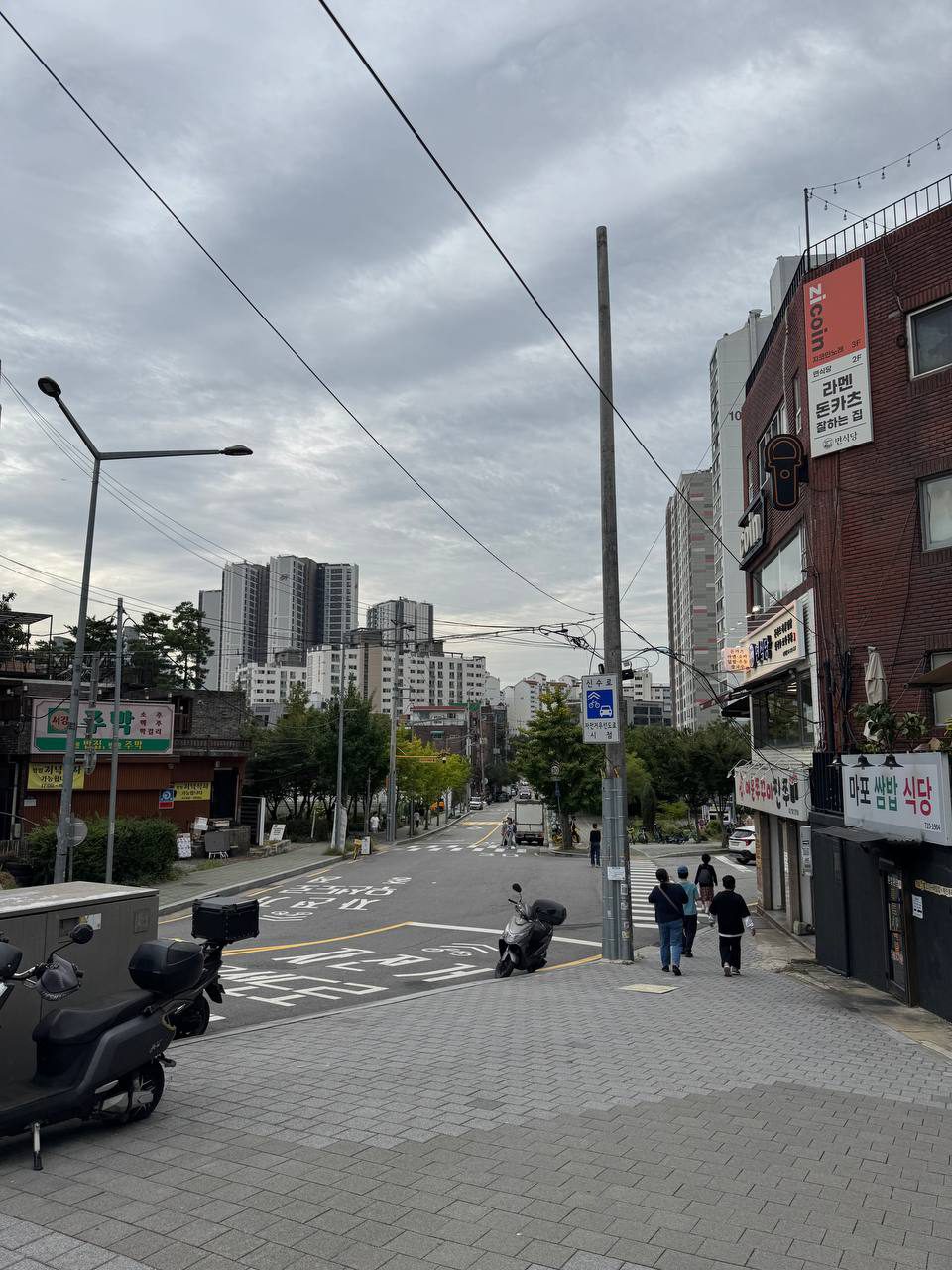 내리막 골목 양옆으로 라멘 돈카츠집, 마포 쌈밥 식당, 주막 간판이 붙어 있고 배달 오토바이가 서 있는 서강대 정문 앞 거리