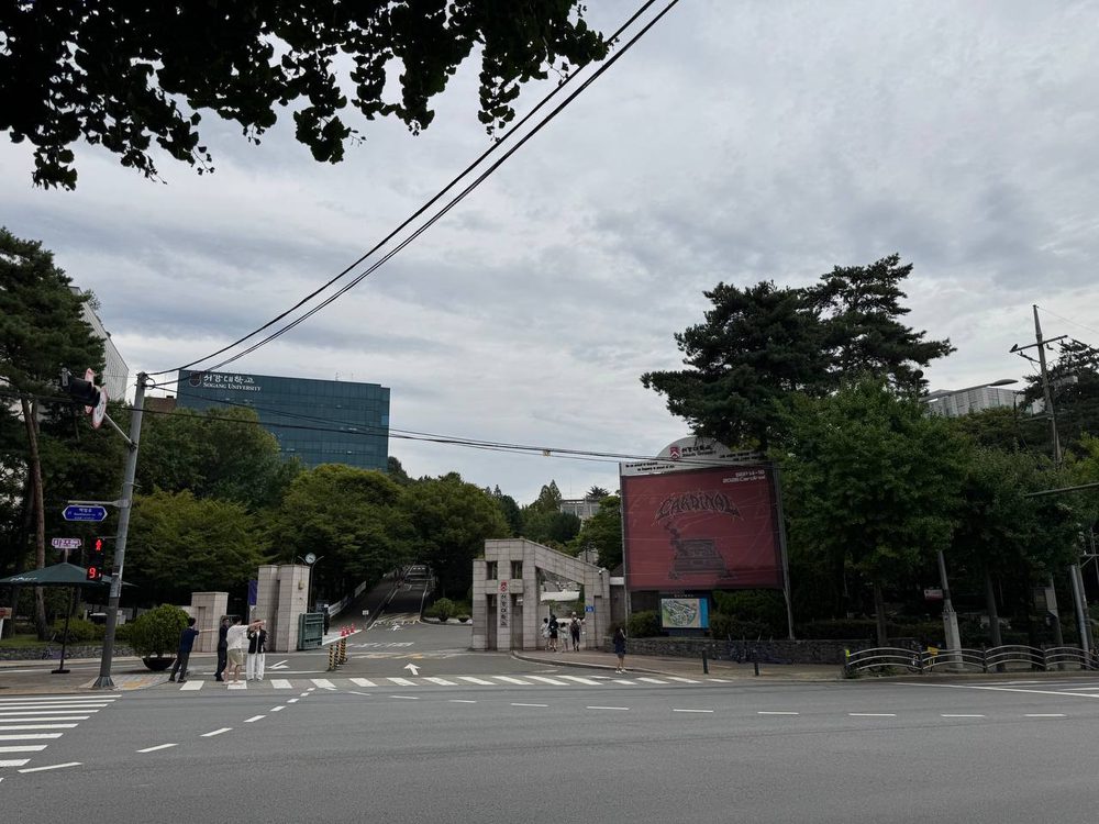 신촌로 건너편에서 본 서강대 정문, 왼쪽에 서강대학교 간판이 붙은 유리 건물과 우거진 나무, 오른쪽에 붉은 공연 포스터 대형 게시판