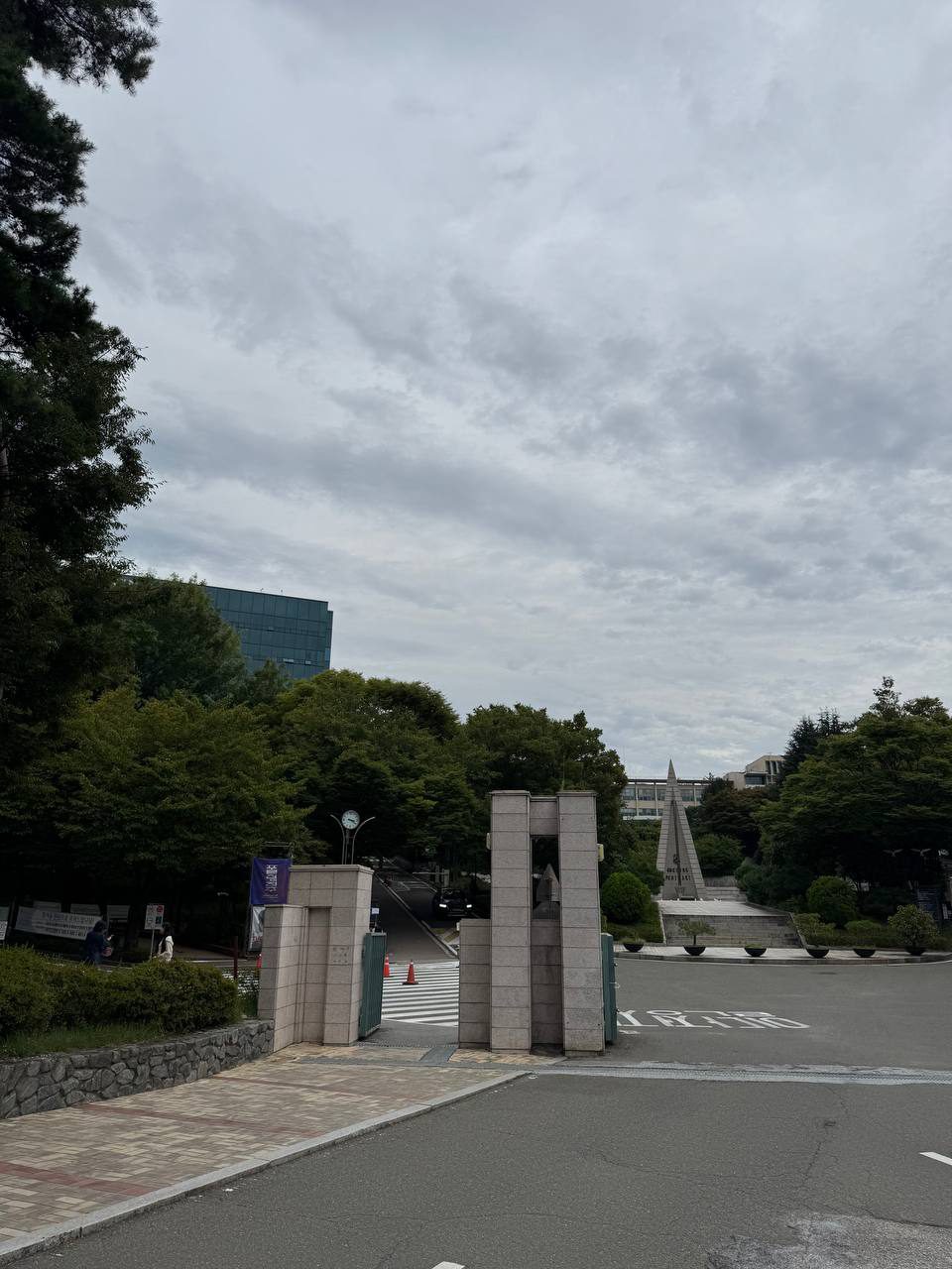 나무 사이로 보이는 알바트로스탑과 정문 기둥, 왼쪽 언덕 위로 유리 건물이 솟아 있다