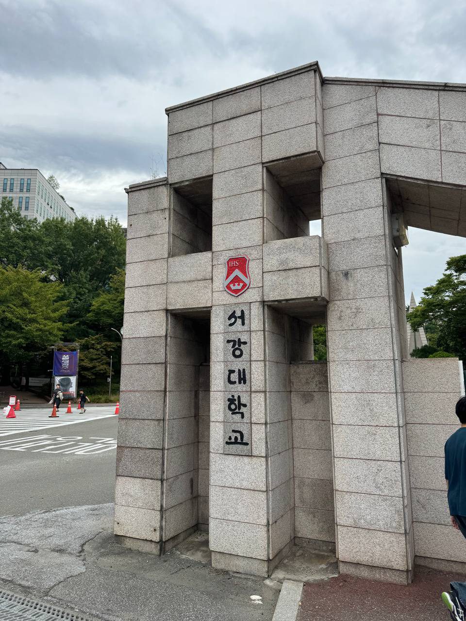 화강암을 쌓아 만든 서강대 정문 기둥, 사각으로 뚫린 구멍과 붉은 IHS 방패 교표, 그 아래 세로로 쓰인 서강대학교 글씨