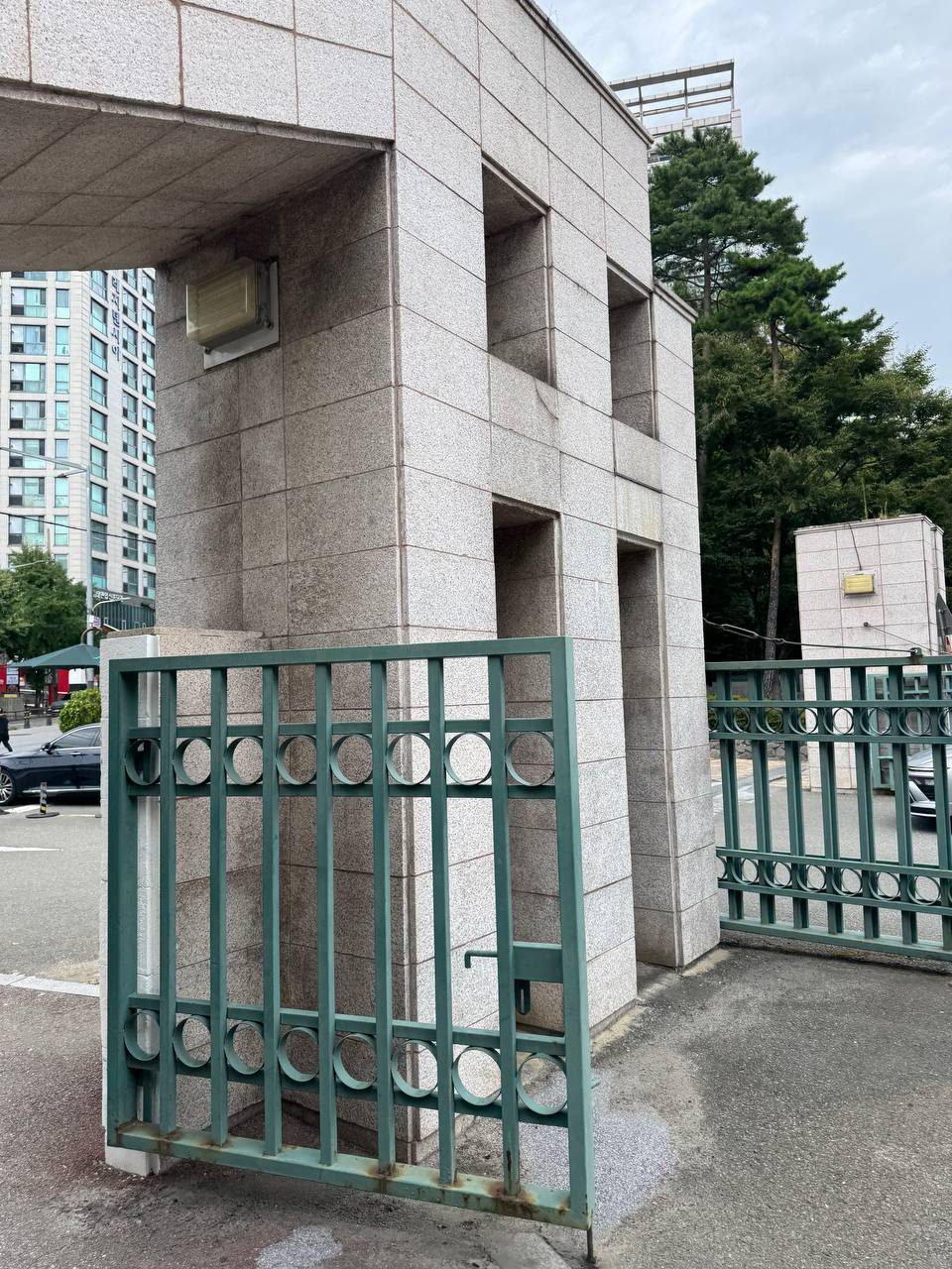 접혀서 기둥에 붙어 있는 청록색 원무늬 철문, 아랫부분에 녹이 번져 있고 뒤로 고층 아파트가 보인다