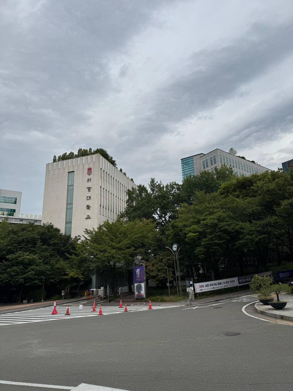 언덕 위로 보이는 흰 건물과 유리 건물, 아래쪽 횡단보도에 라바콘이 늘어서 있고 현수막이 걸려 있다