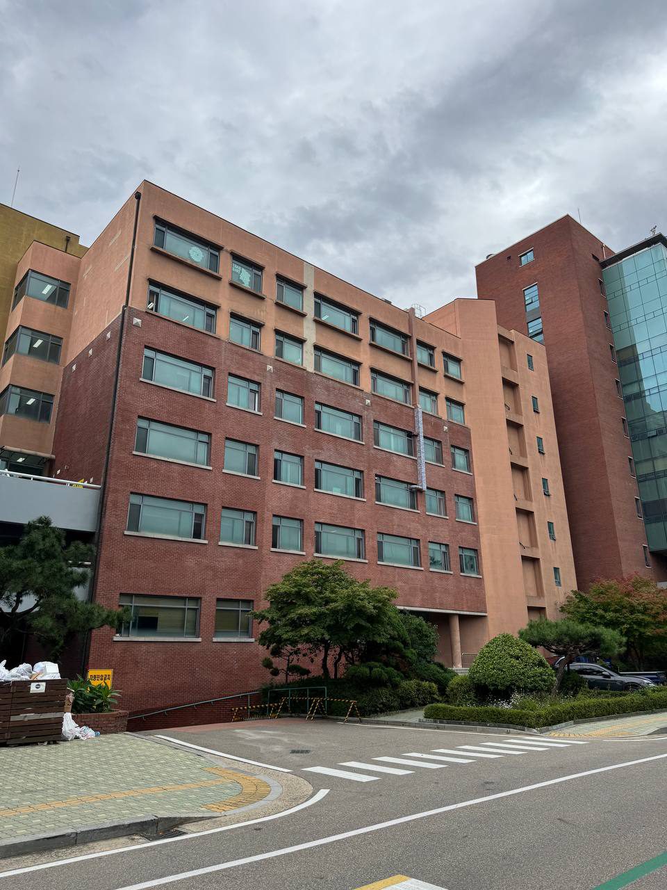 붉은 벽돌과 살구색 외벽의 건물에 창문이 줄지어 있고, 앞쪽 화단과 횡단보도 위로 흐린 하늘이 펼쳐져 있다