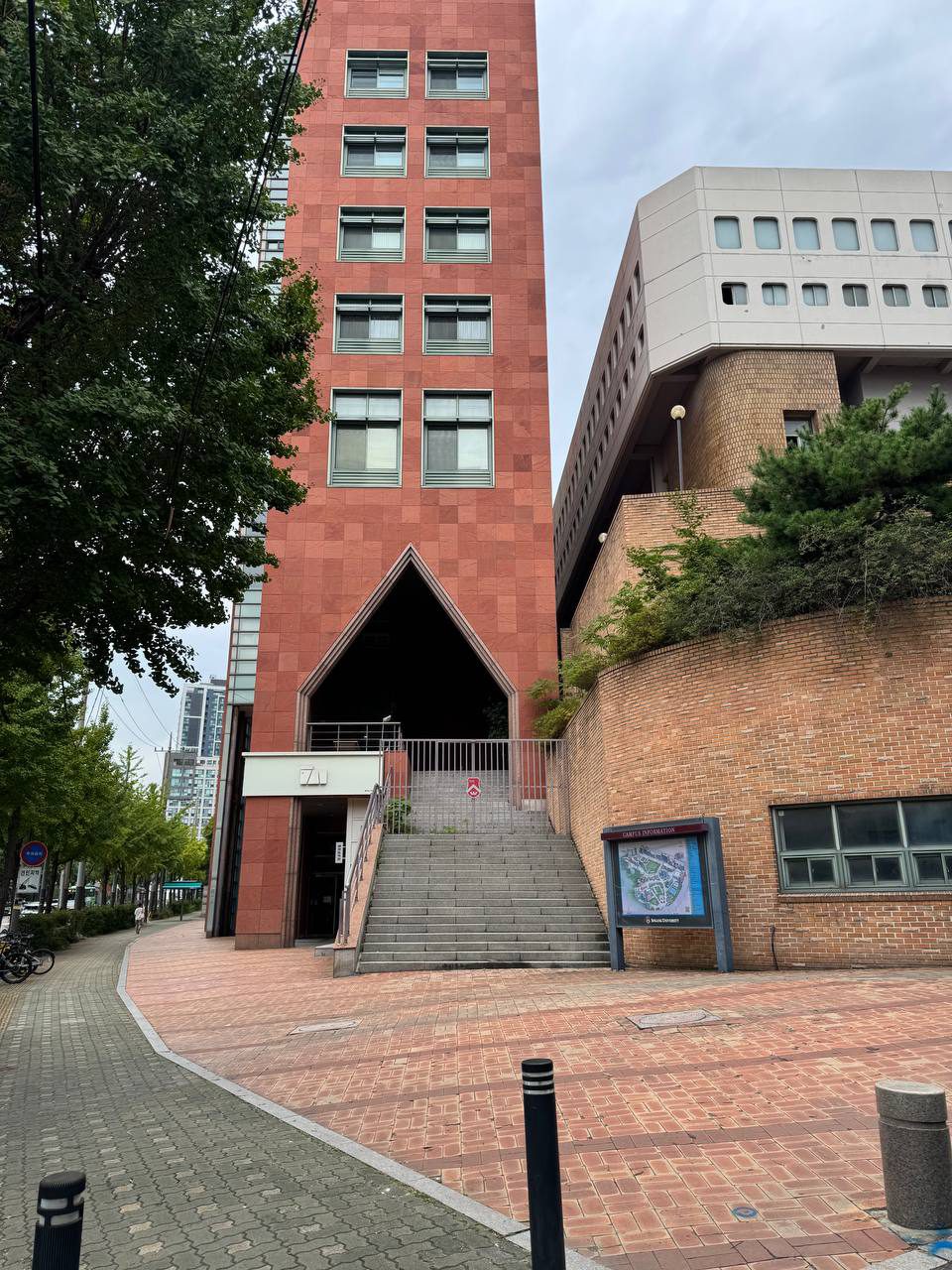붉은 석재로 마감된 높은 동문회관 건물과 삼각형 아치 아래 계단, 오른쪽에 둥근 창이 있는 건물과 캠퍼스 안내도