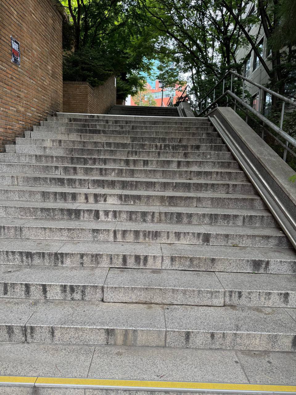 붉은 벽돌 옹벽을 끼고 위로 길게 이어지는 화강암 계단, 계단 끝 너머로 나무와 캠퍼스 건물이 보인다
