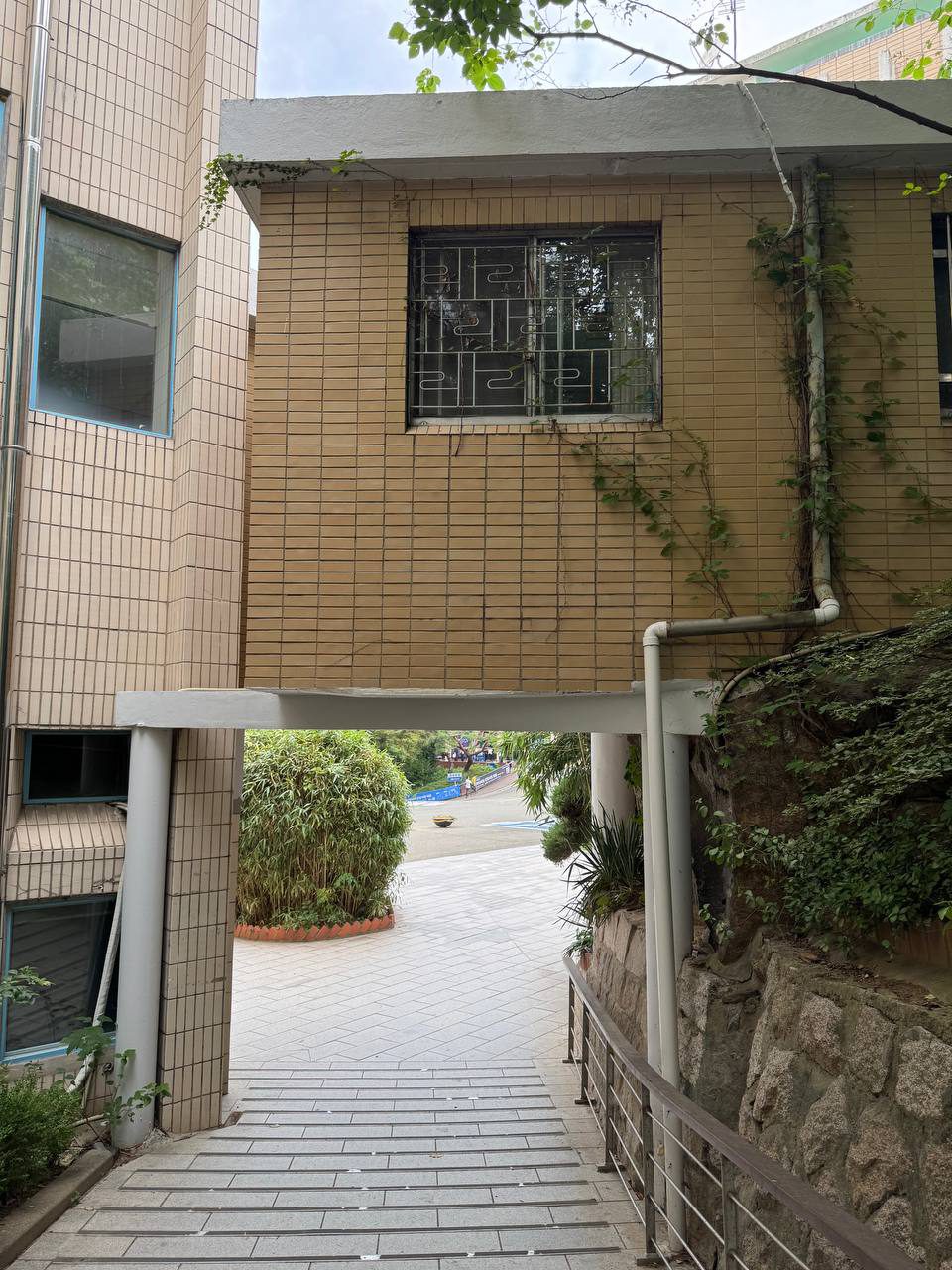 베이지색 타일 건물 아래로 계단길이 이어지고, 오른쪽 바위에는 덩굴이 자라 있는 통로 전경