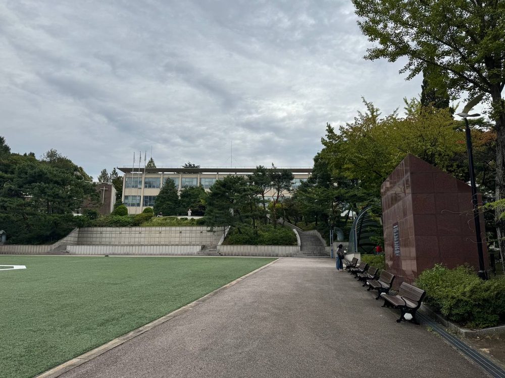 광장 옆 산책로를 따라 벤치들이 이어지고, 붉은 기념비와 가로등 너머로 본관 건물과 소나무가 보인다