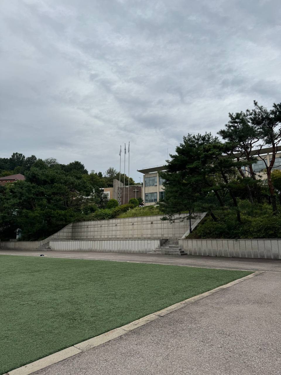 인조잔디 너머 층층이 쌓인 석축과 계단, 그 위로 소나무와 깃대 세 개, 십자가가 달린 건물과 본관이 보인다