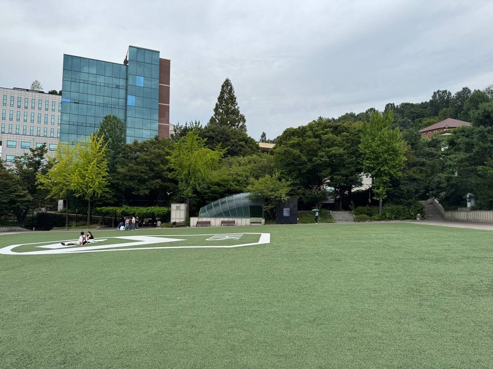 반대편에서 본 청년광장, 흰 로고가 그려진 인조잔디 위에 두 사람이 앉아 있고 뒤로 유리 외벽 건물과 유리 돔 구조물, 나무 그늘 아래 사람들이 보인다